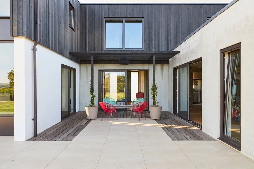 Modern outdoor patio with sliding glass doors, potted plants, a round table, and colorful chairs.