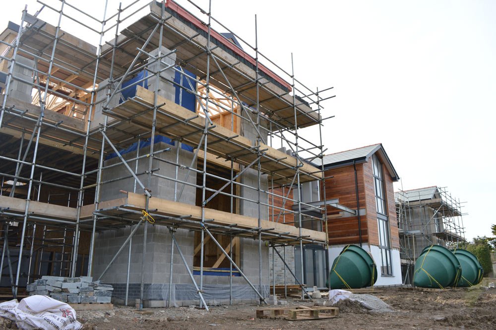 Construction site with scaffolding around a multi-story house under construction, with wooden and concrete elements and green waste containers outside.