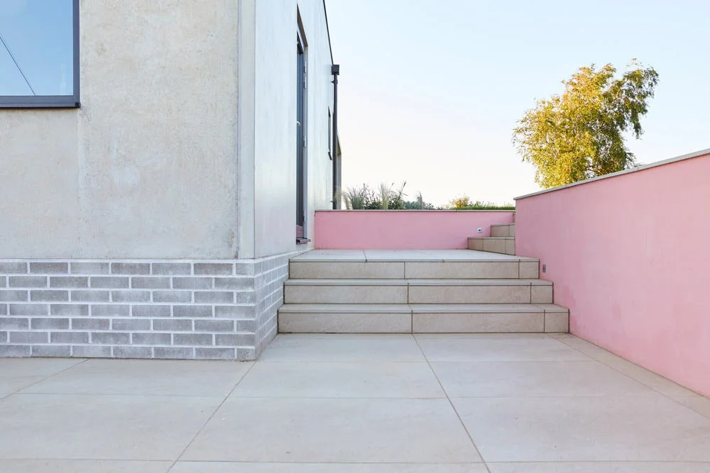Exterior of a modern building with stairs, a pink wall, and a background tree with yellow and green leaves.