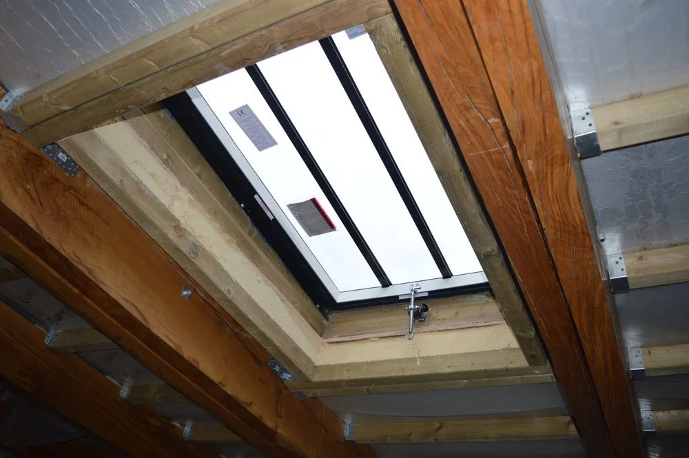 Construction site with a ceiling opening fitted with a skylight window, surrounded by wooden framing and metal brackets.