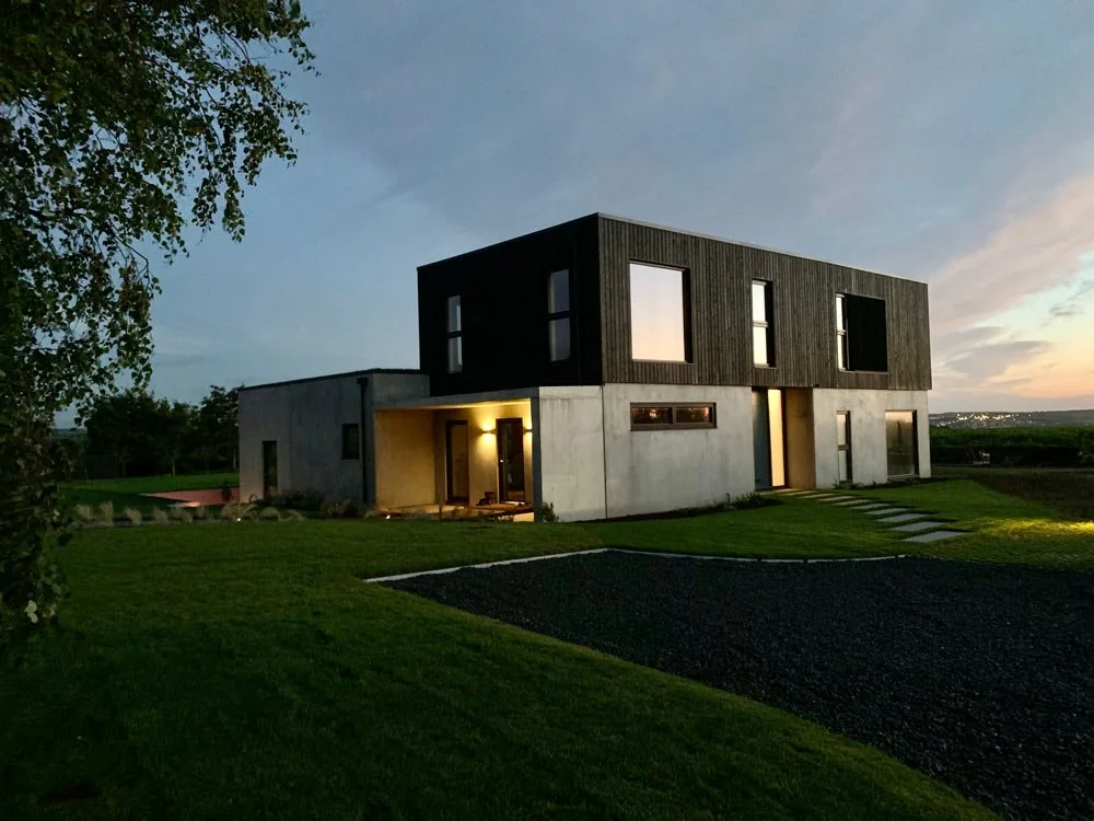 Modern two-story house with concrete and dark wood exterior, surrounded by green lawn under evening sky.