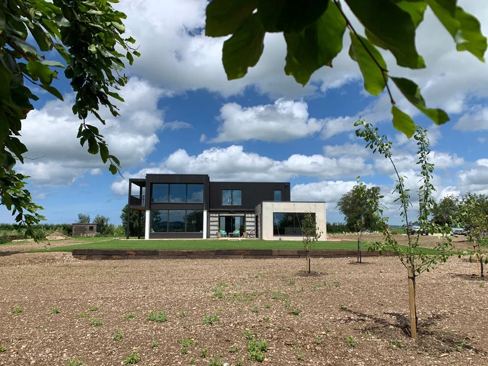 Modern two-story house with large glass windows, located in a rural area with young trees and a cleared yard, under a partly cloudy sky.