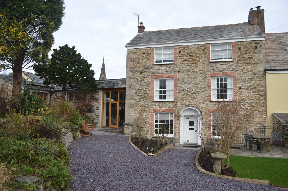 A three-story stone house with white window frames and a white front door. The house has a garden with a curved gravel pathway, a wooden bench, and some small trees.