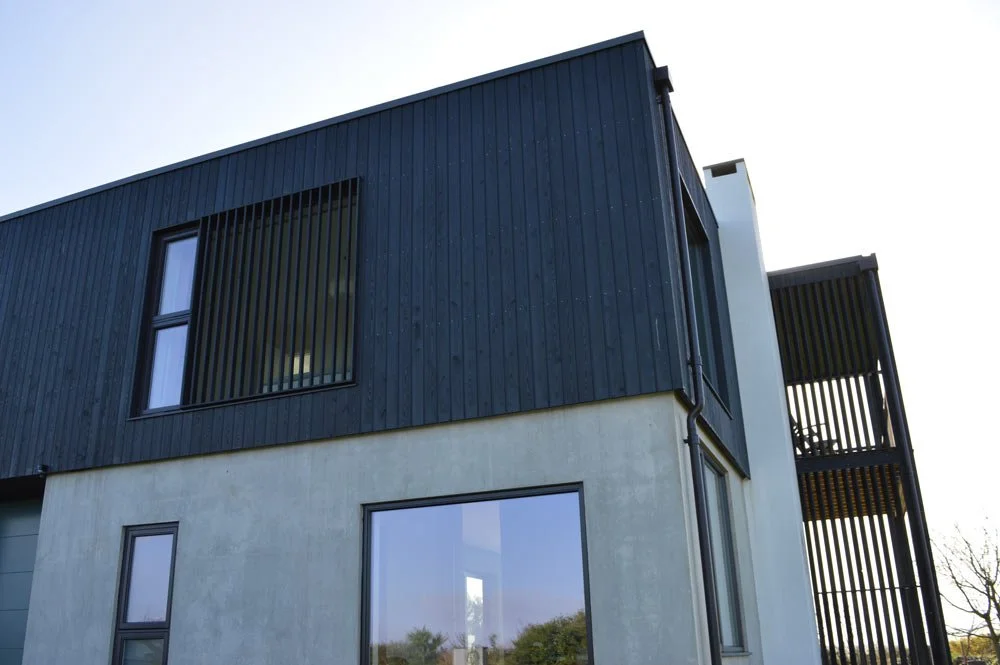 Modern two-story house with dark vertical wood siding on upper level and light concrete on lower level, large windows, and a balcony with vertical slats.