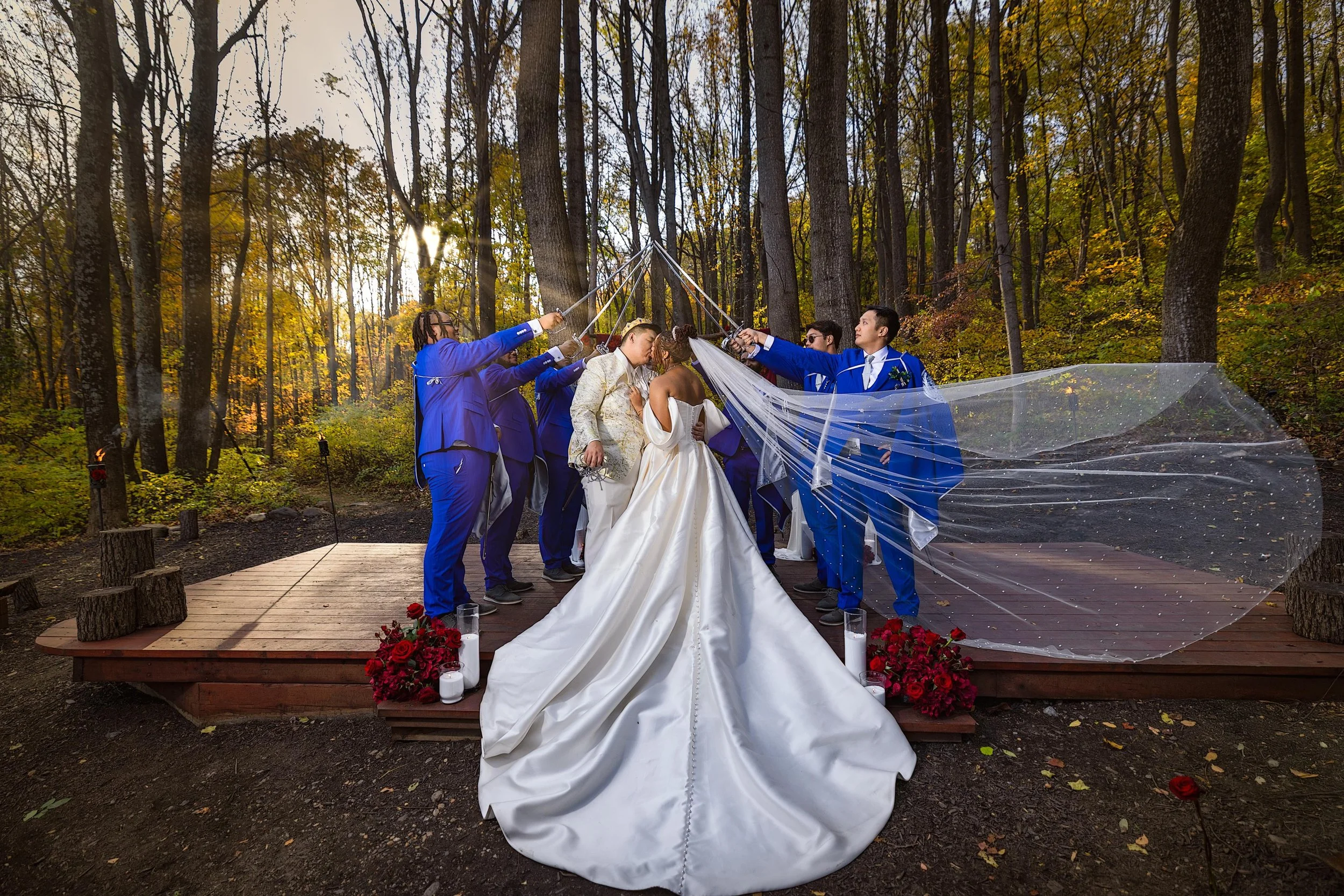 Bride and groom in wedding attire standing on a wooden platform surrounded by bridesmaids and groomsmen, in a forest setting during sunset, with a veil flowing in the wind and red roses decorating the scene.