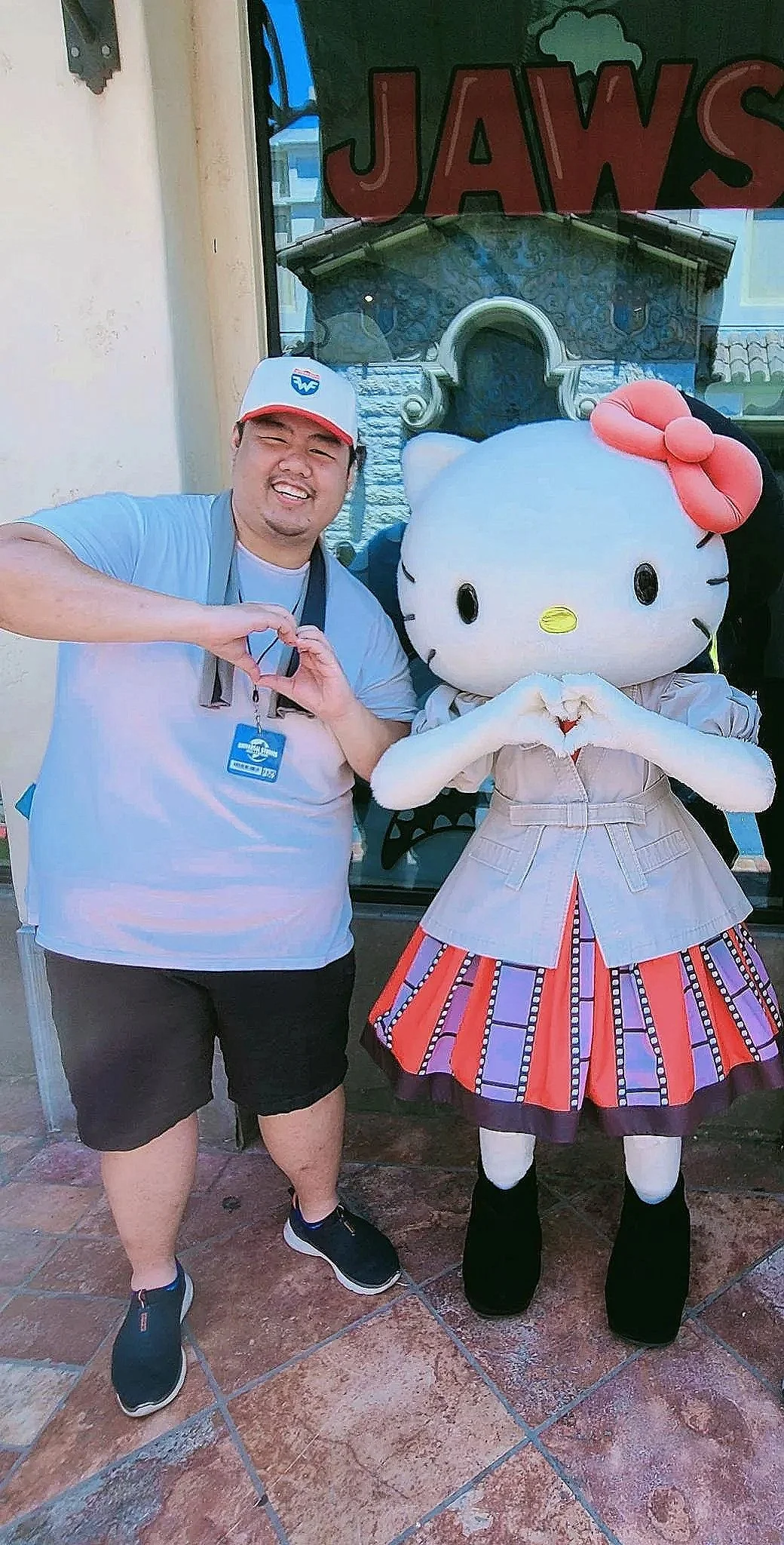 A man and a person in a Hello Kitty costume making a heart hand gesture in front of a store window.