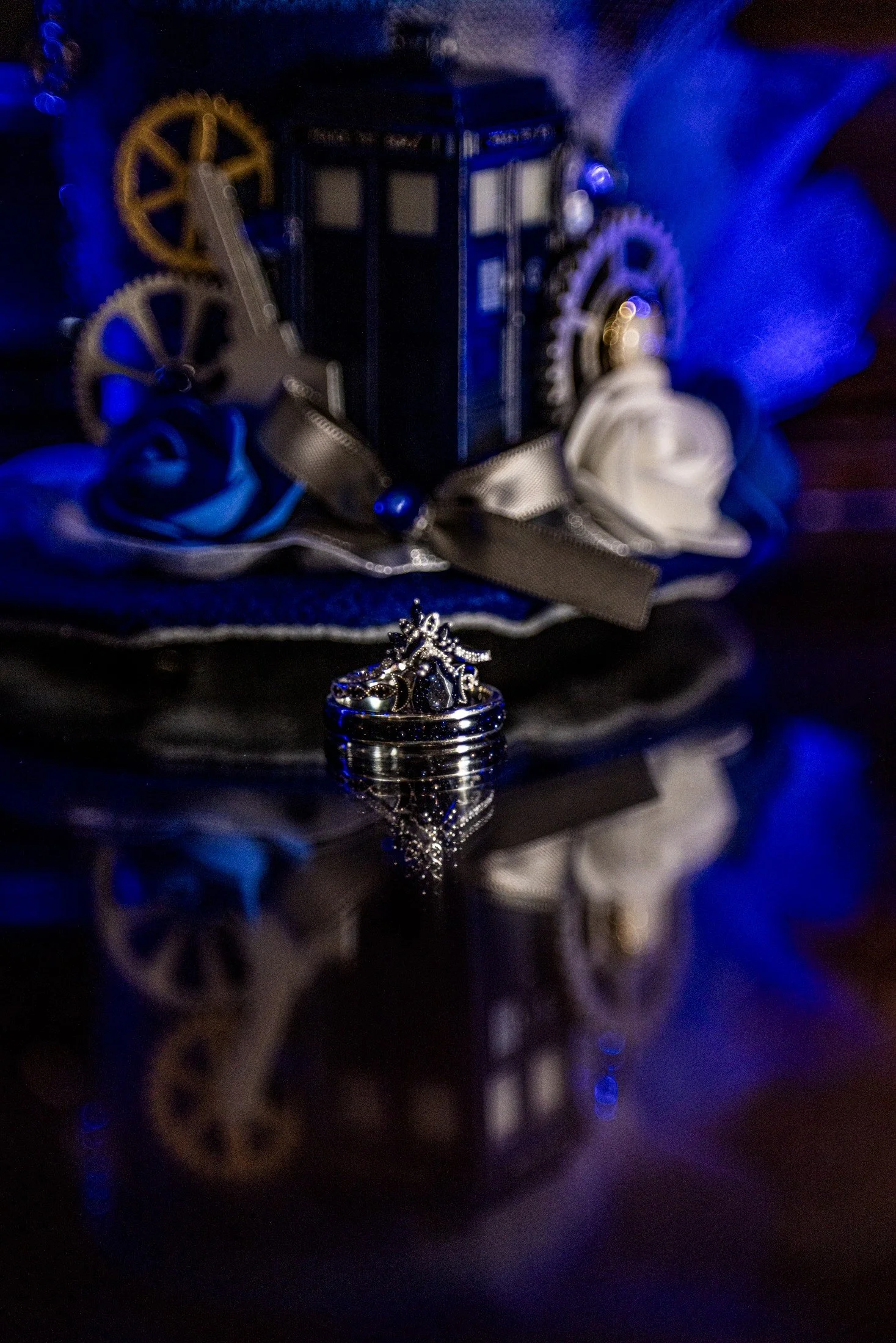 A close-up of a silver ring with a gemstone, reflecting on a glossy surface. In the background, there is a dark decorative box with gears and ribbons, with blue and white fabric and accessories around it.