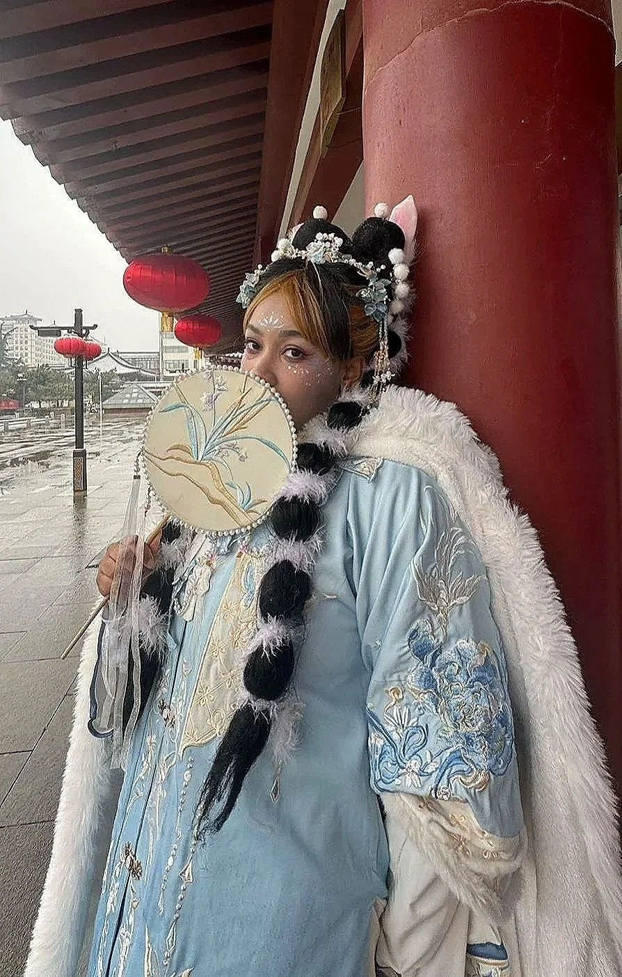 Person in traditional Chinese costume holding a decorated fan, standing under a red lantern canopy on a rainy day.