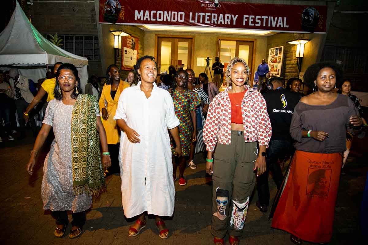 Group of women walking and smiling in front of a building with a banner that reads 'Macondo Literary Festival.' The women are dressed in colorful and varied clothing, and some are wearing wristbands. There are tents and other people in the background
