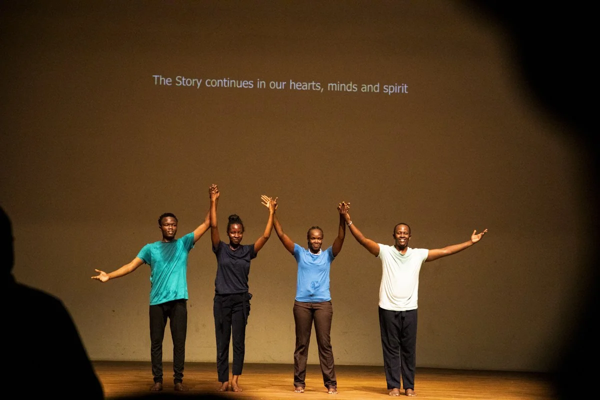Four people standing on stage with their hands raised and holding hands, smiling. Text on the screen behind them reads, "The Story continues in our hearts, minds and spirit."