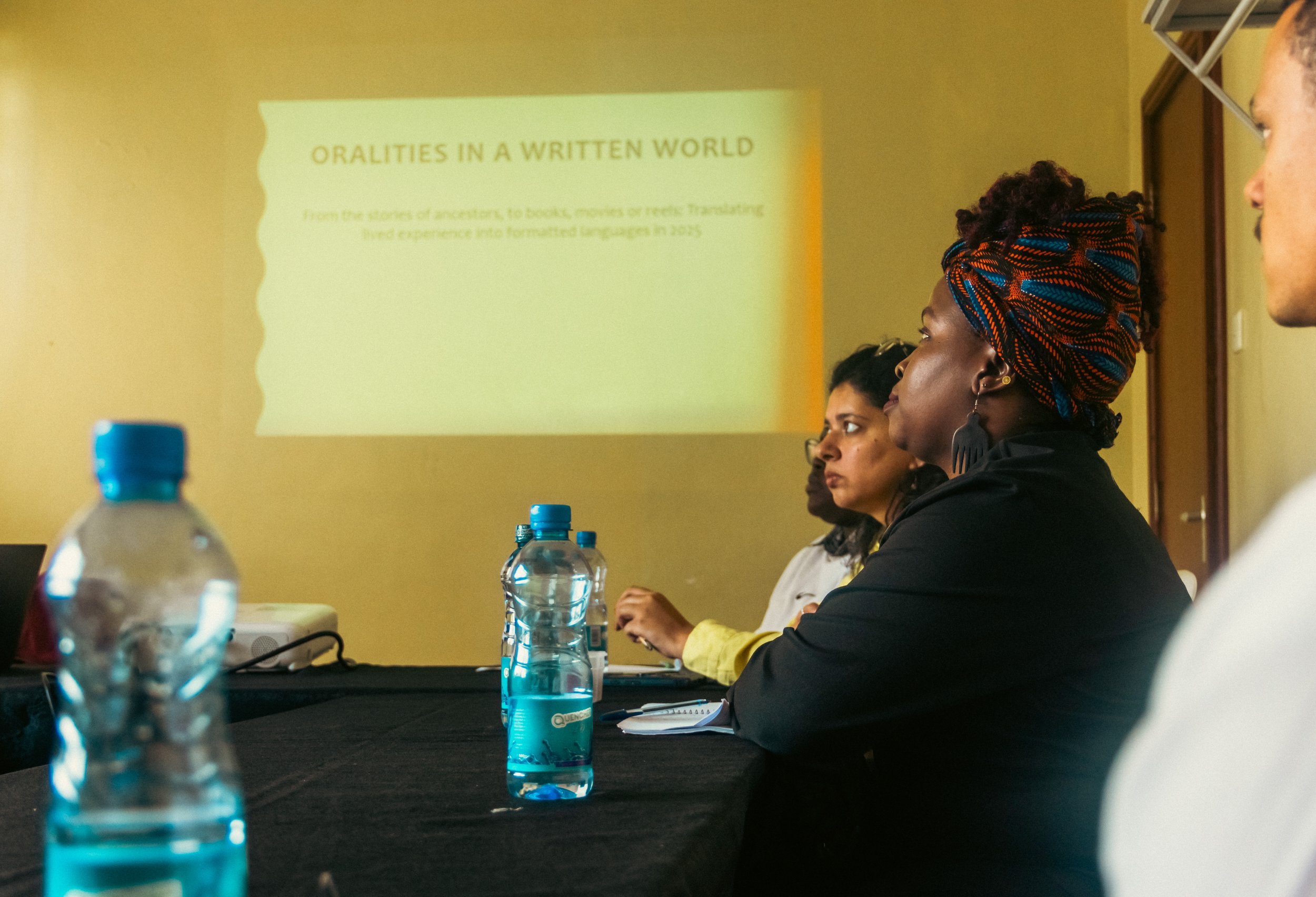Group of people sitting at a conference table with water bottles, attending a presentation with a slide projected on the wall titled 'Oralities in a Written World.'