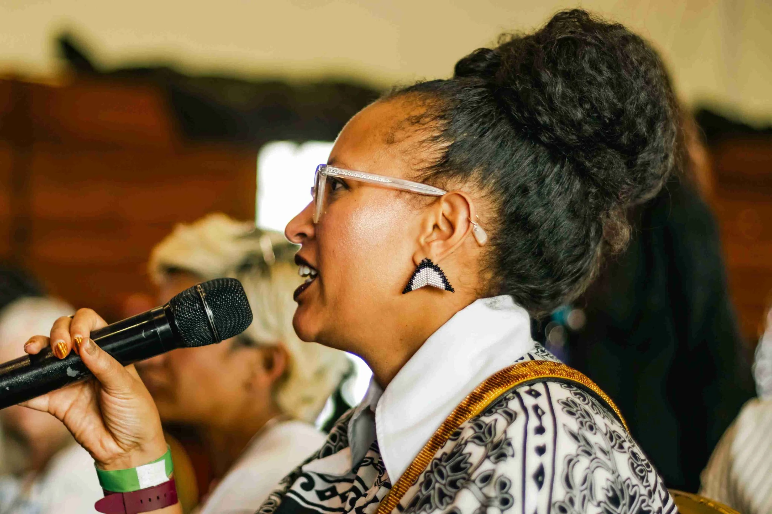 A woman with glasses and large earrings speaking into a microphone during a conference or event.