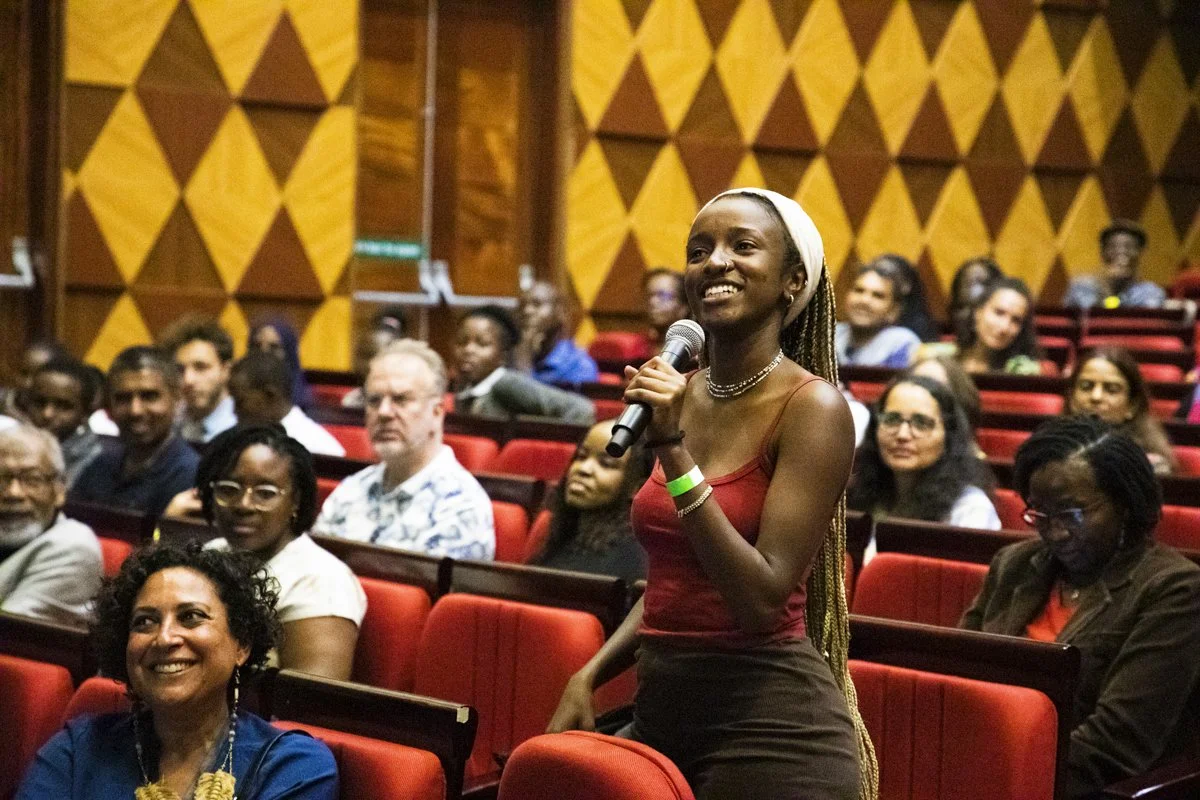A woman with a white headscarf is standing in an auditorium, smiling, and holding a microphone. The audience around her is seated, paying attention, and some are smiling. The auditorium has red seats and wood-paneled walls.