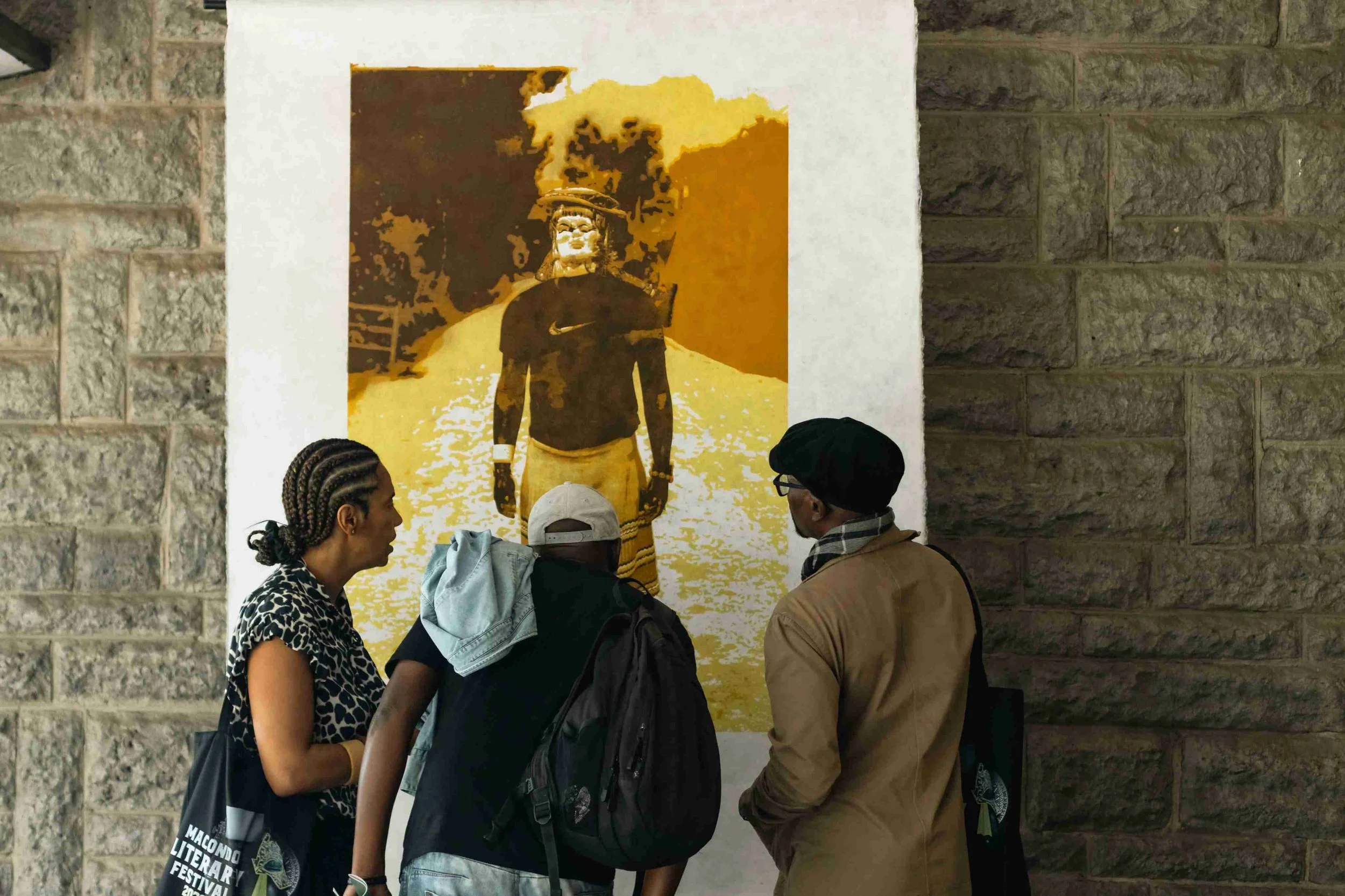 Three people looking at a large poster or photograph of a person in a hat, outdoors, with trees and a pathway in the background.