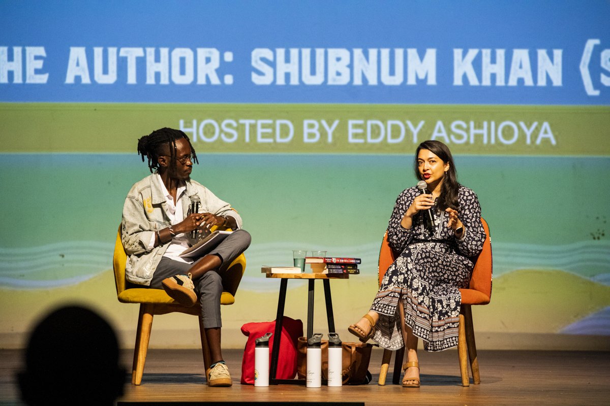 A woman in a patterned black and white dress and a woman in a casual jacket and pants sit on orange chairs on stage, engaging in a discussion as part of a book event. They each hold a microphone, and a small table between them holds books and water b