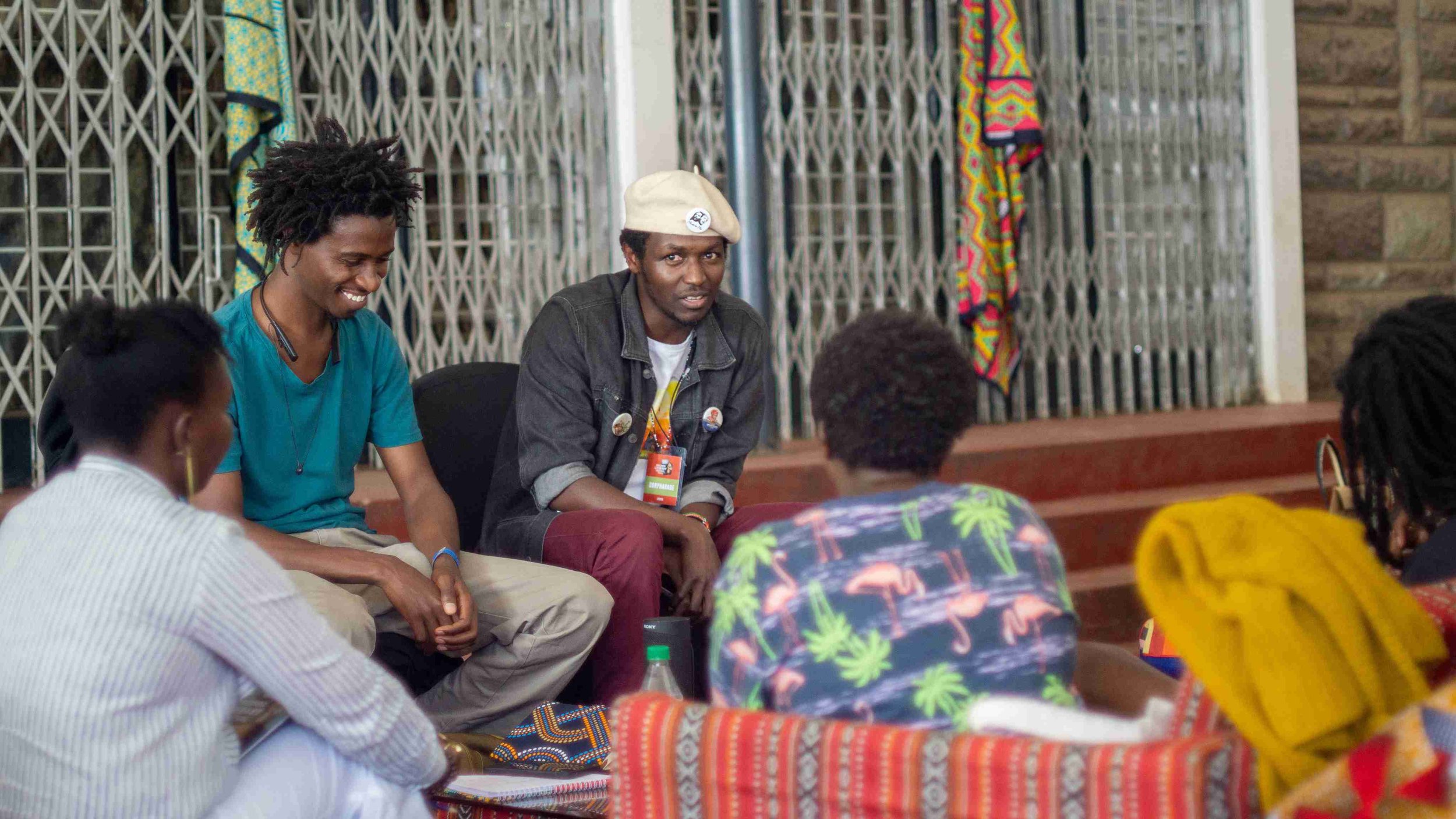 A group of people sitting in a circle having a discussion indoors. Two individuals in the foreground, one wearing a white shirt with a colorful pattern and the other in a bright yellow scarf, face away from the camera. A woman with a big smile, weari