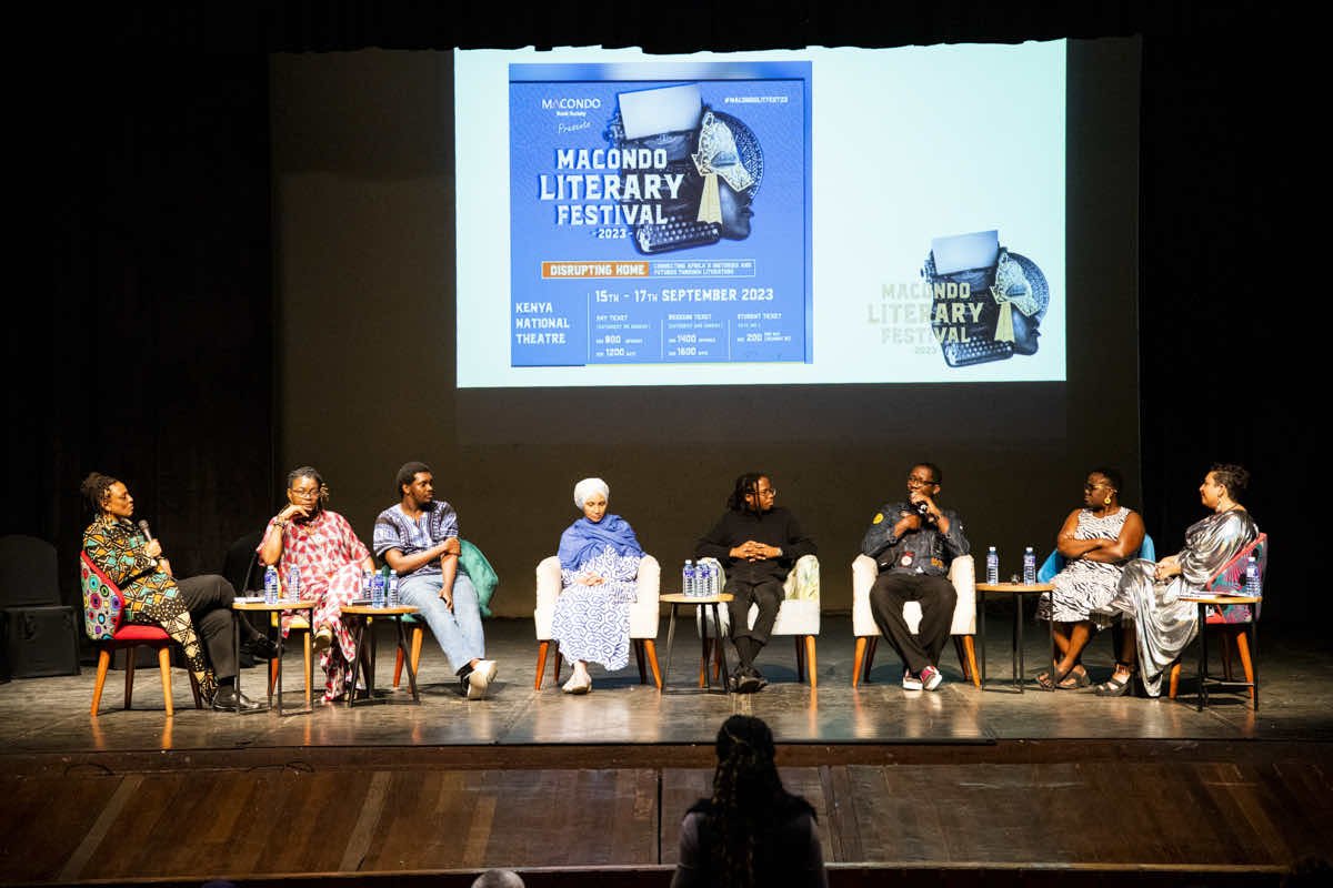 Panel discussion at the Macondo Literary Festival 2023, with seven diverse speakers seated on stage, some holding microphones, with a large festival poster projected behind them on a black background.