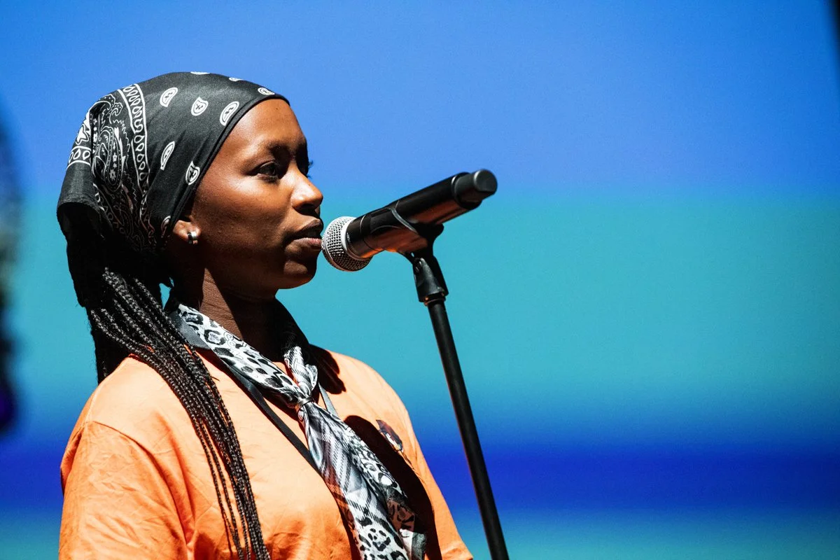 A woman with long braided hair, wearing a black bandana, speaking into a microphone against a blurred blue background.