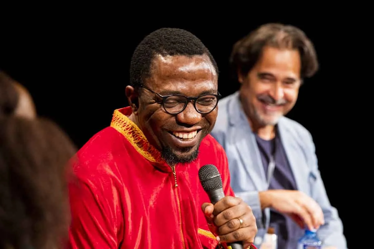 Two men on stage, one in a red shirt and glasses holding a microphone, smiling, and another man in a light blue shirt with long hair smiling, against a black background.