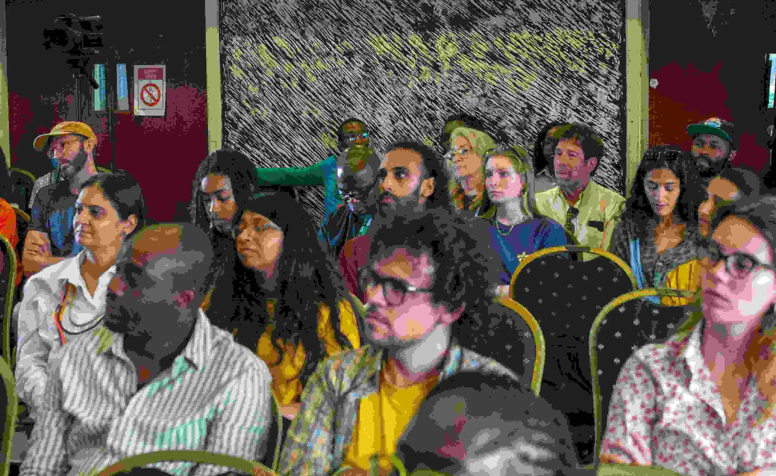 A group of diverse people attending an indoor event, seated in chairs, some wearing glasses, with a colorful wall background.