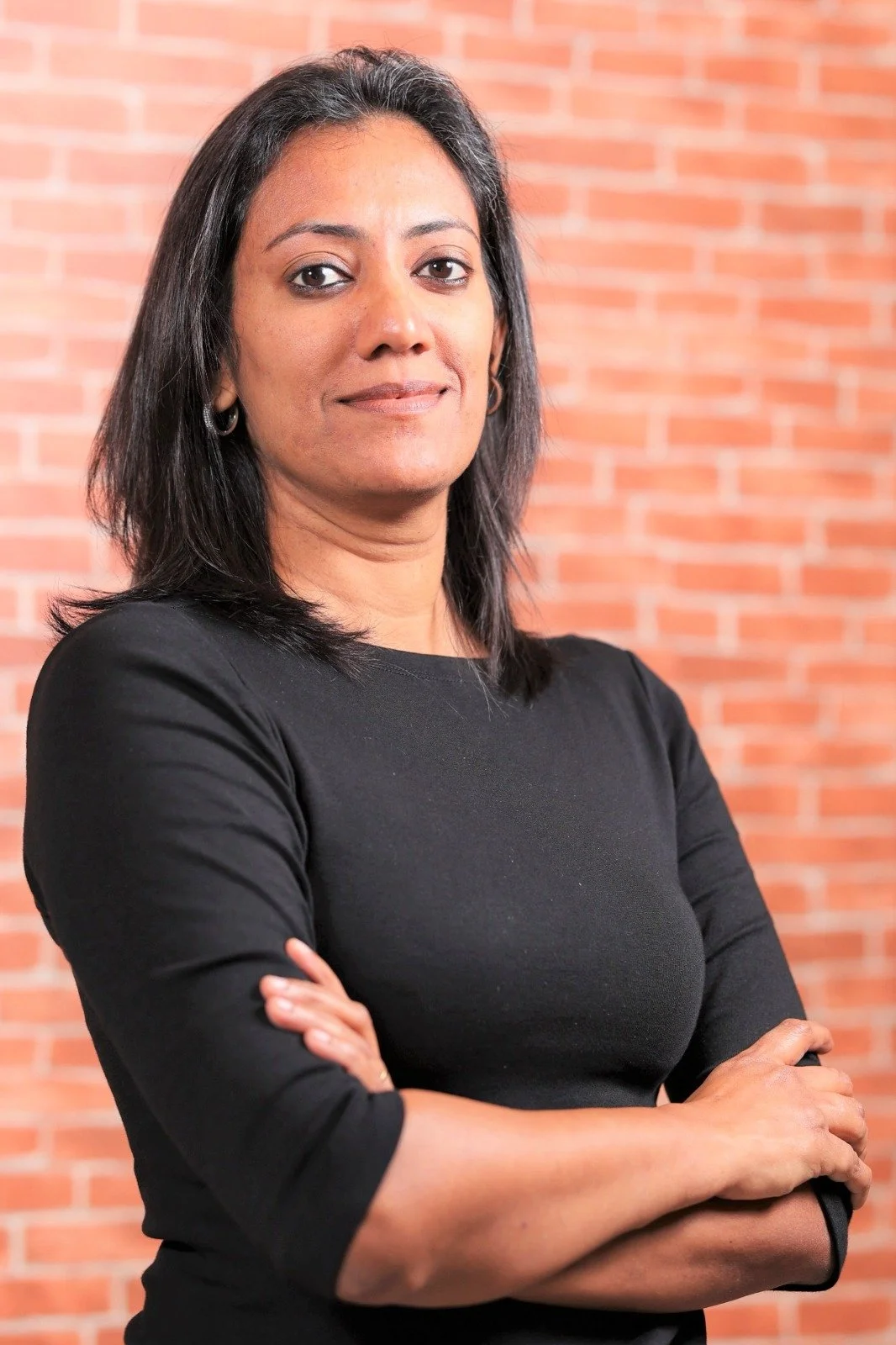 A confident woman with shoulder-length black hair, wearing a black top, standing with her arms crossed in front of a red brick wall.