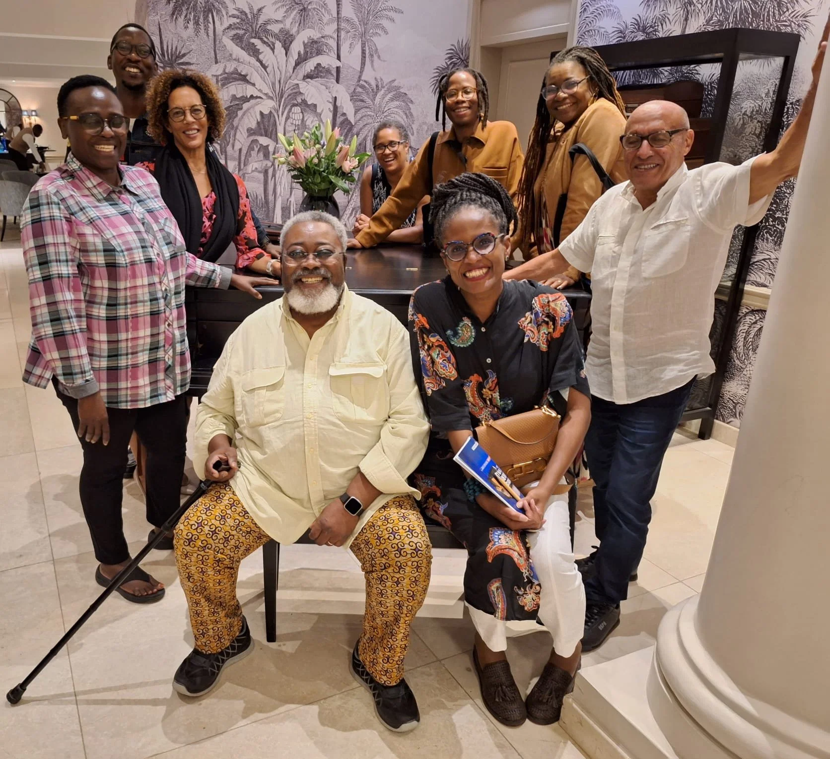 Group of smiling people posing together indoors, with a floral wallpaper background, a table with flowers, and a column visible.