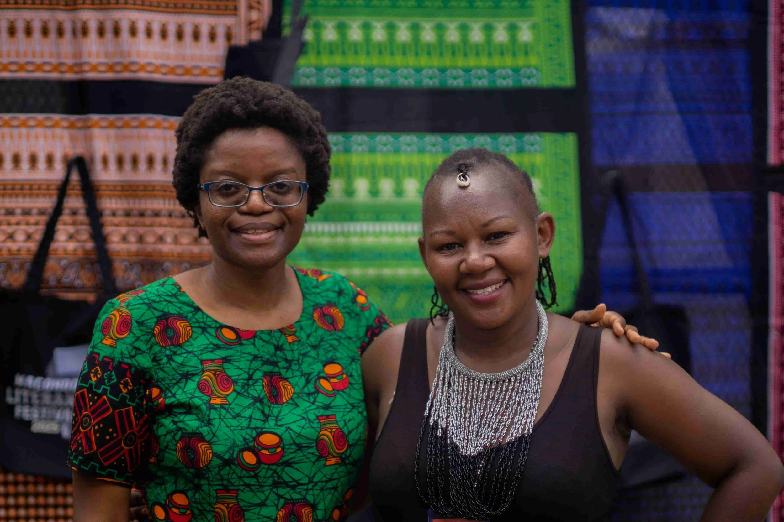Two women standing together, smiling at the camera, with colorful patterned textiles displayed in the background.