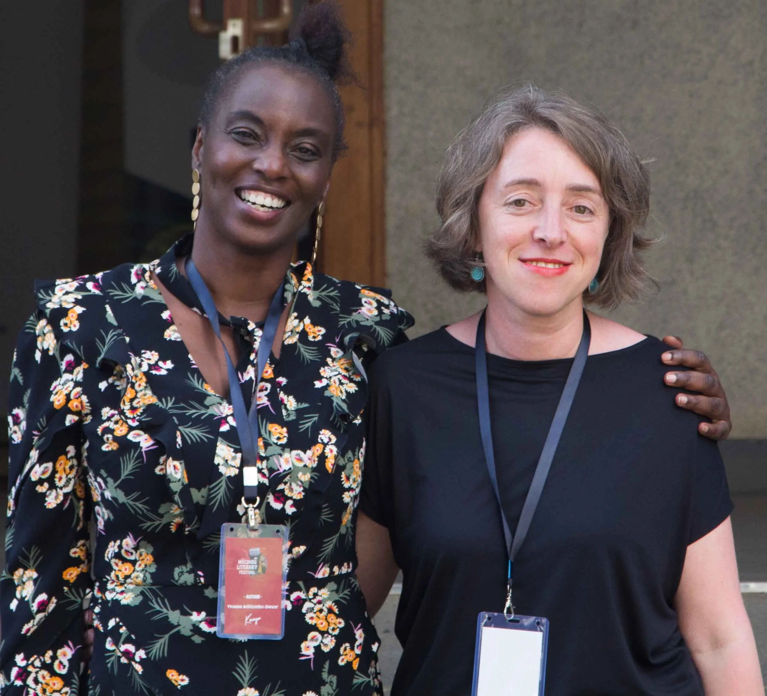 Two women standing outdoors, smiling at the camera. One woman has dark skin and is wearing a black floral dress with earrings, and the other has light skin and is wearing a black shirt with turquoise earrings. They both have conference badges around their necks.