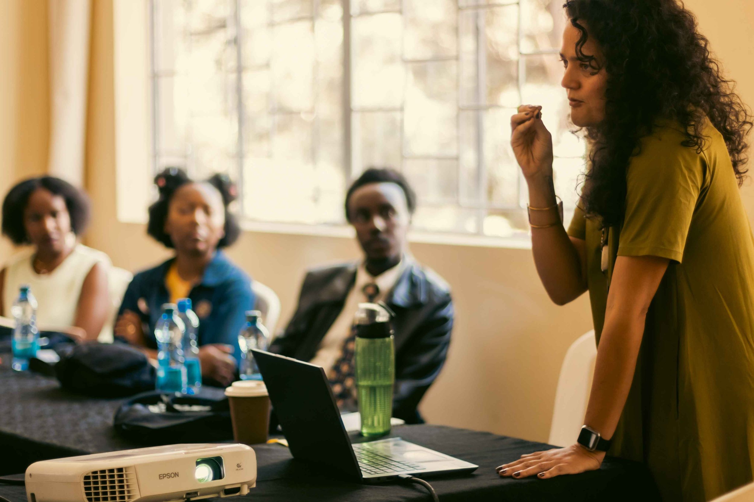 A woman standing and speaking to a group of three women in a meeting room. The standing woman is wearing a green dress, has curly hair, and is gesturing with her right hand. The women sitting at the table have various hairstyles and are listening att