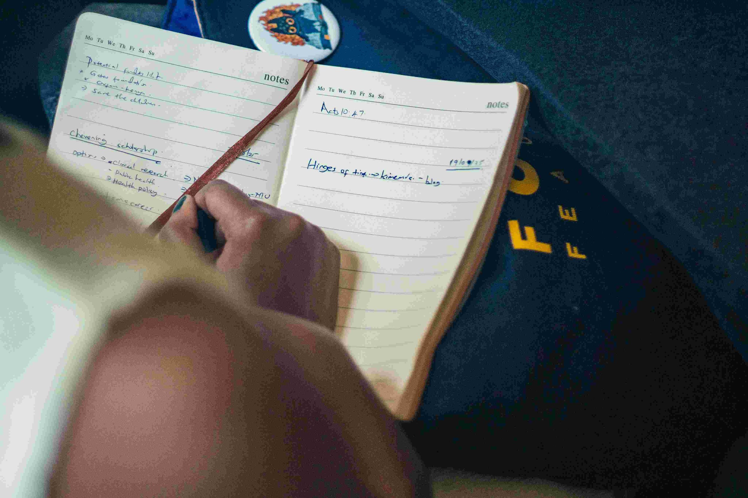 An open notebook with handwritten notes on the left page and a date on the right page, resting on a dark fabric surface with some yellow letters and a sticker of a tree on a circular background nearby. A person is writing in the notebook with a black