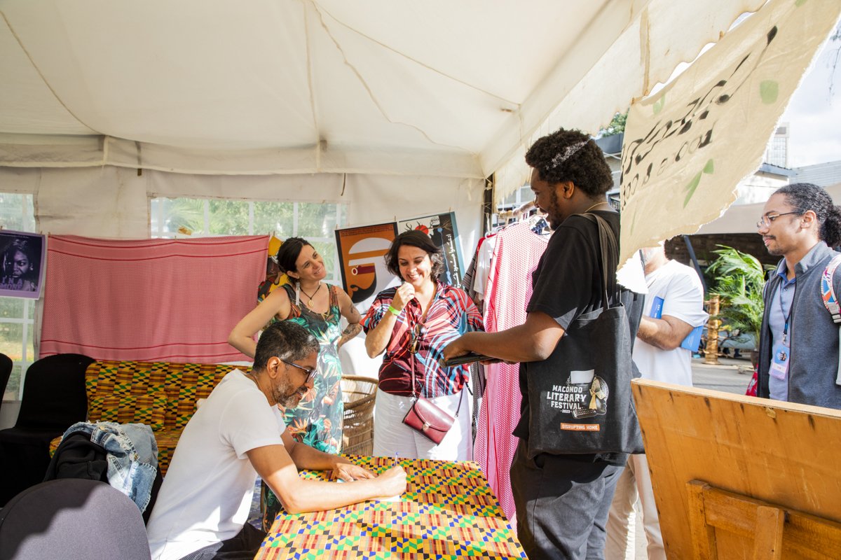 A group of people at a literary festival under a tent, engaging in conversation at a table with colorful cloth, with artwork and clothing displays in the background.