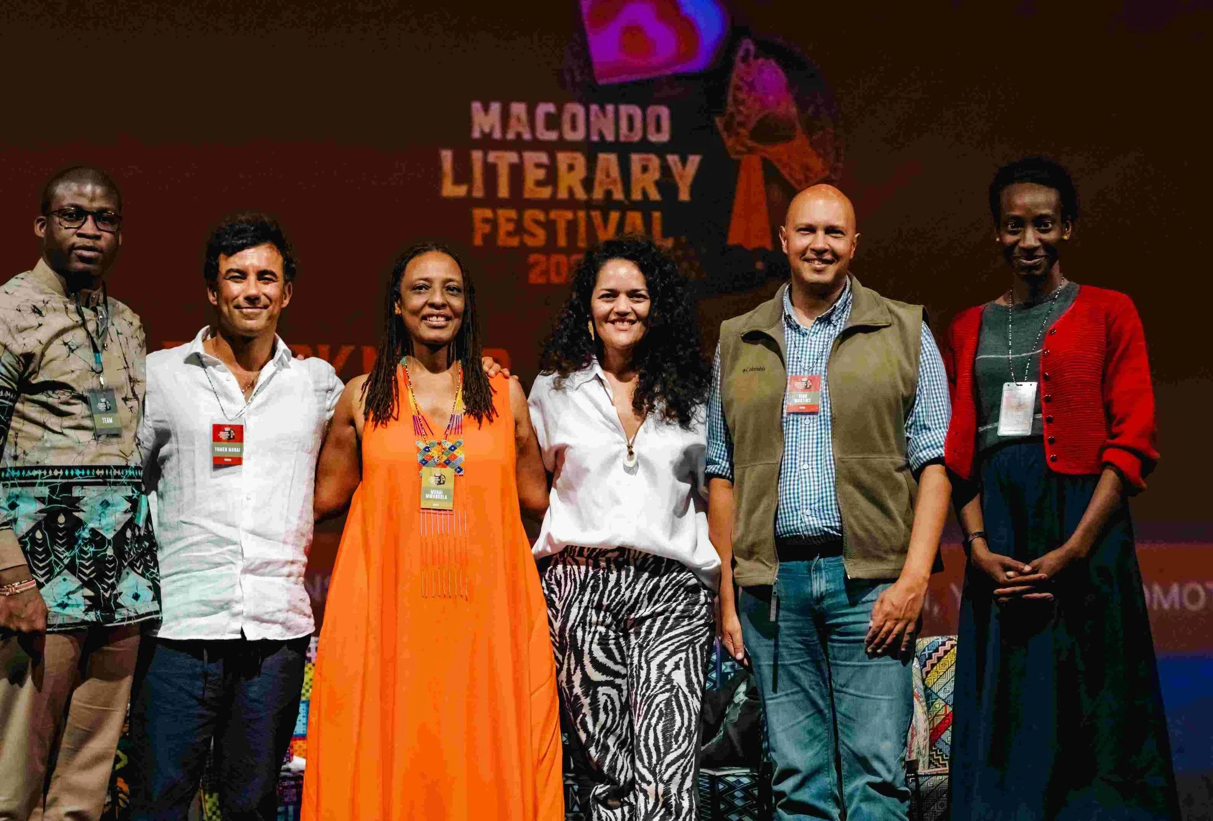 Group of seven diverse people standing together on stage at Macondo Literary Festival, with festival banner in background.