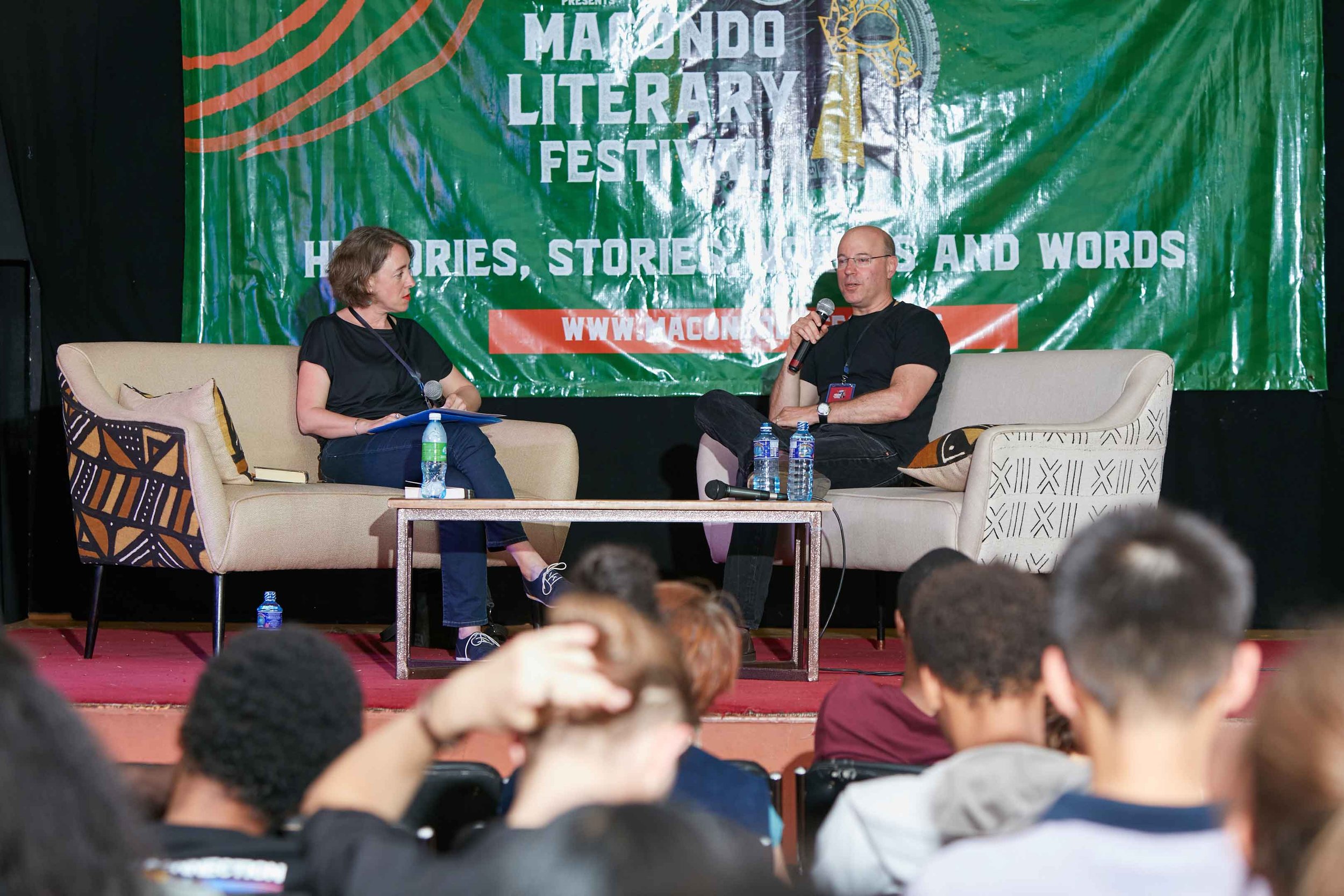 A woman and a man seated on stage at the Mangundo Literary Festival, engaging in a discussion. The woman holds a microphone, and the man is speaking into a microphone. A table with water bottles in front of them, and a large green and orange banner w