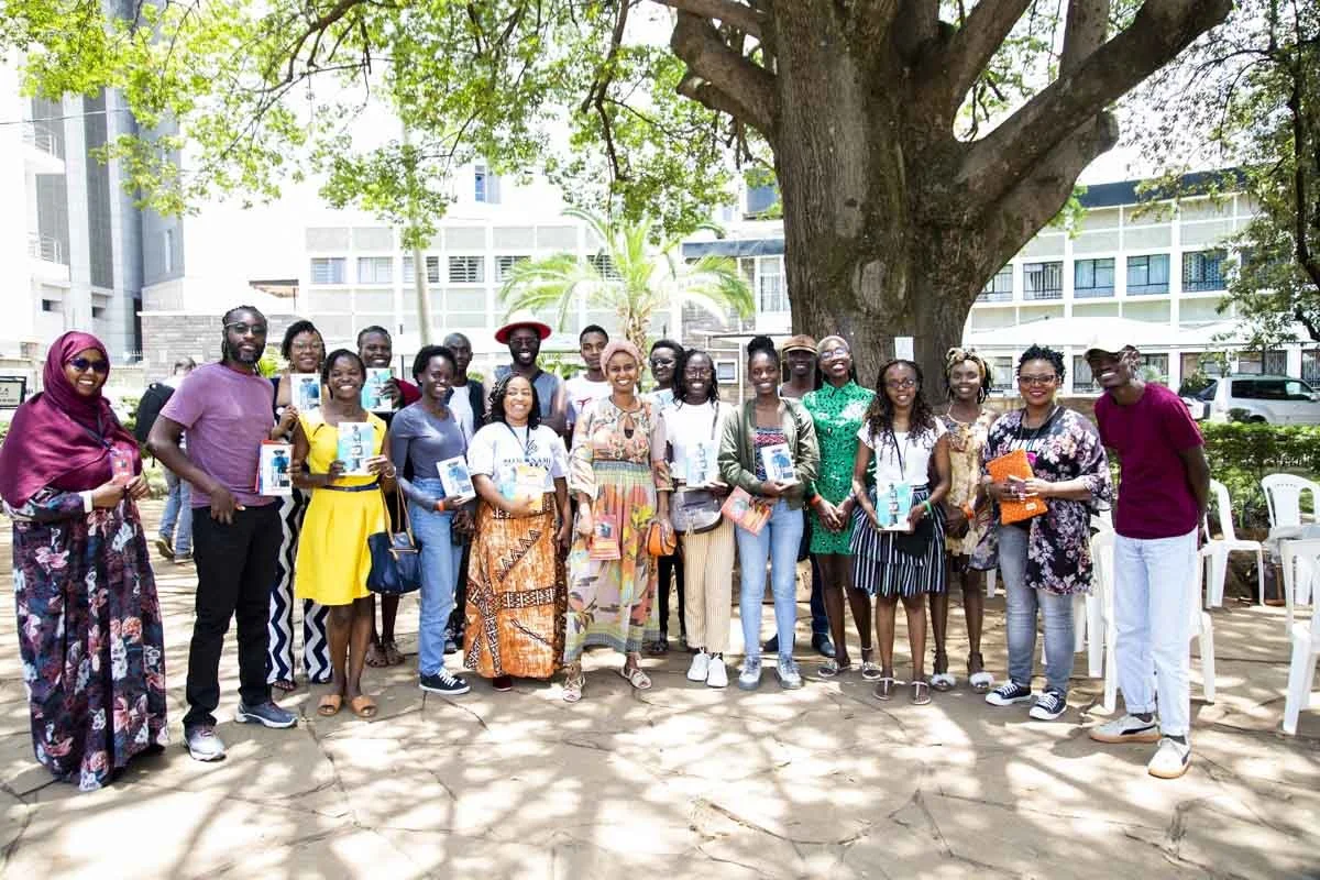 A group of people gathered outdoors under a large tree, holding books or pamphlets, smiling for a photo during daytime in an urban setting.