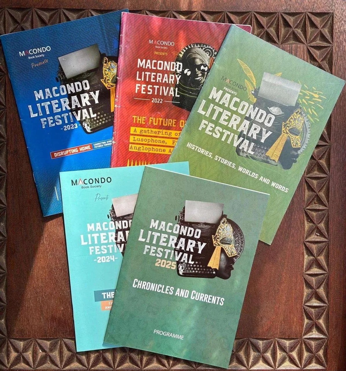 Five conference programs for the Macondo Literary Festival from 2023 to 2025, each with colorful covers featuring a typewriter, a mask, and cultural artwork, laid out on a wooden table.