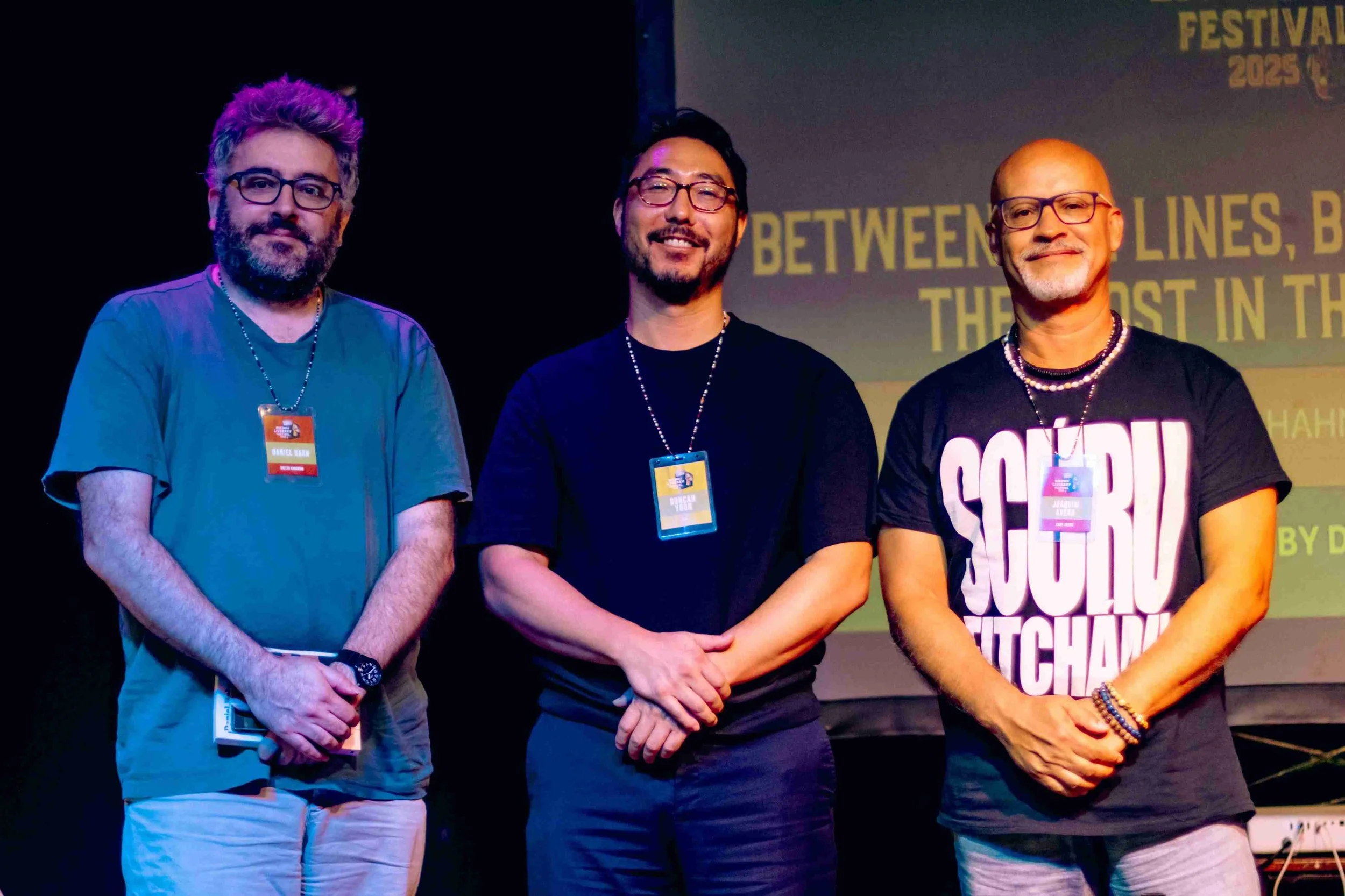 Three men standing on stage with a screen behind them displaying text about a festival in 2025. The man on the left has curly, purple-tinted hair, a beard, and glasses, wearing a green T-shirt. The man in the middle has short dark hair, glasses, and 