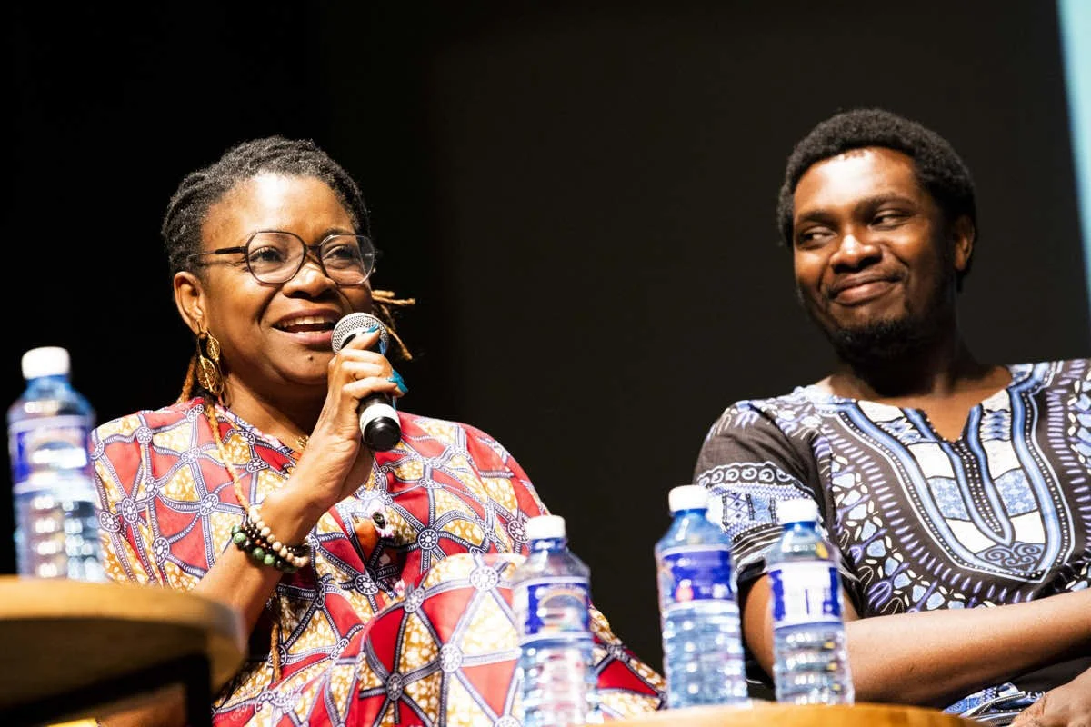 A woman with glasses and earrings speaking into a microphone, smiling, seated next to a man with a beard and short hair, also smiling, at a panel with water bottles on the table.