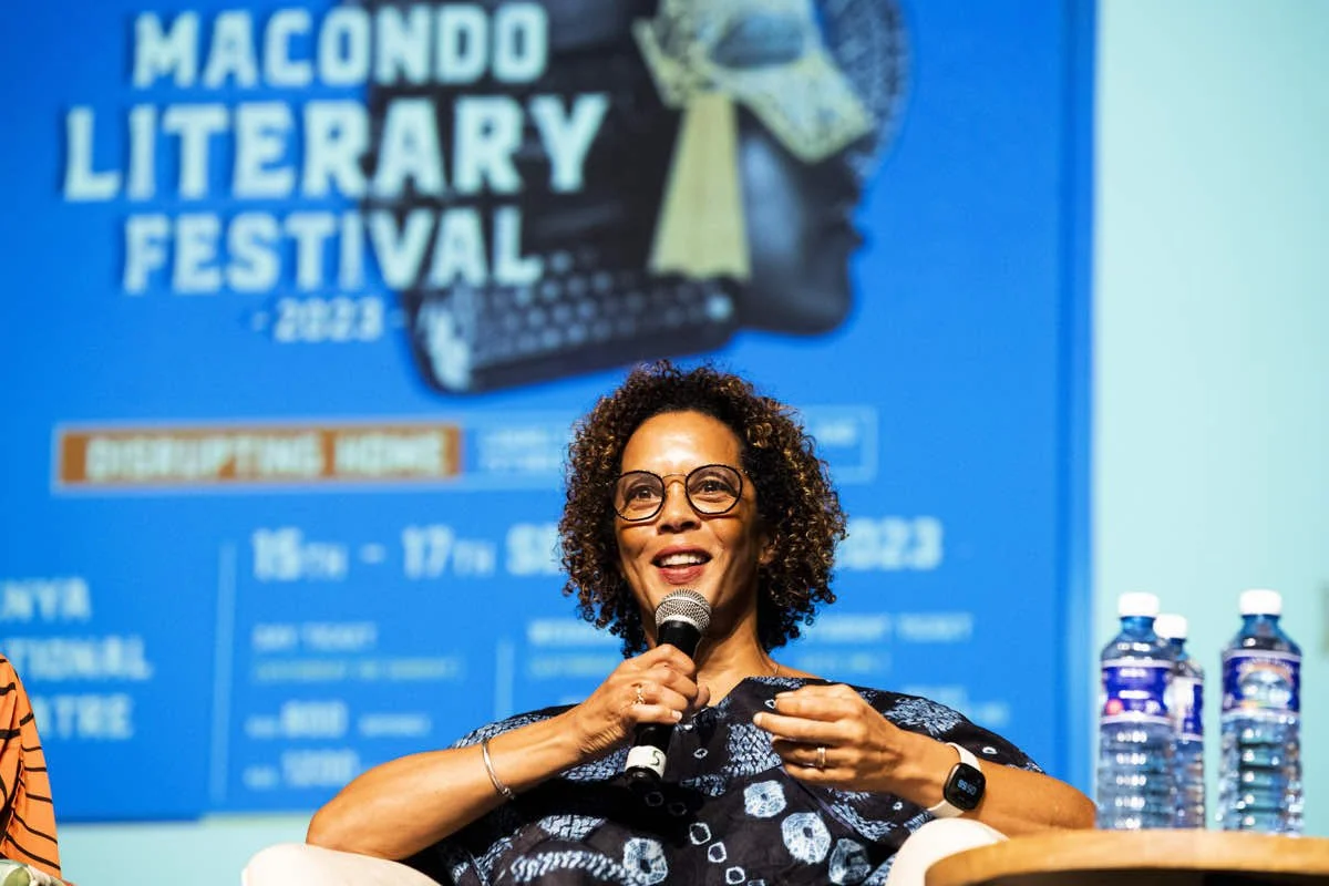 A woman with curly hair and glasses speaking into a microphone at the Macondo Literary Festival 2023, with a blue festival poster in the background and water bottles on the table.