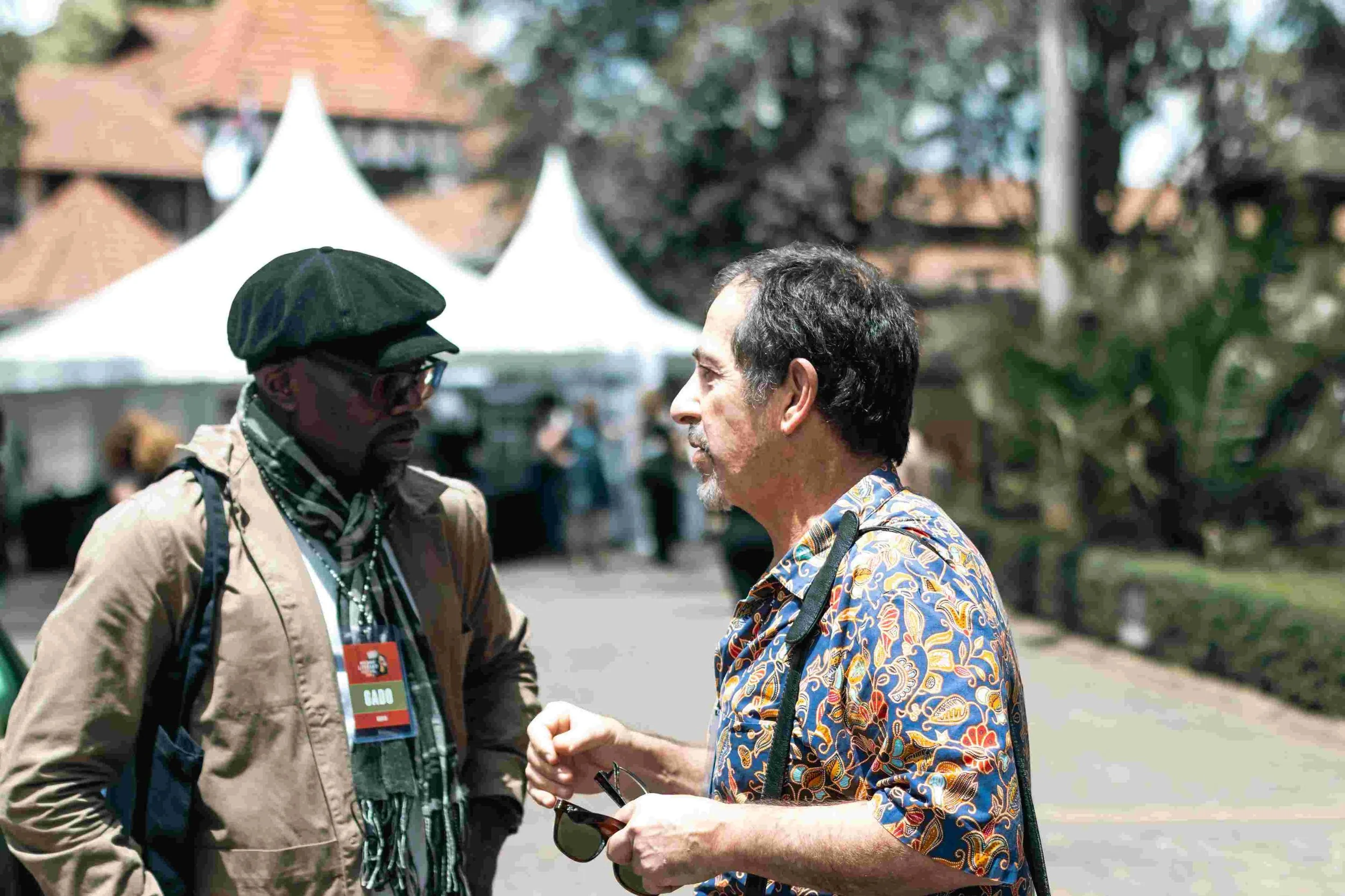 Two men talking outdoors at an event, one wearing a black cap, glasses, a tan jacket, and a badge, the other with curly dark hair and a colorful short-sleeve shirt, holding sunglasses, with tents and trees in the background.