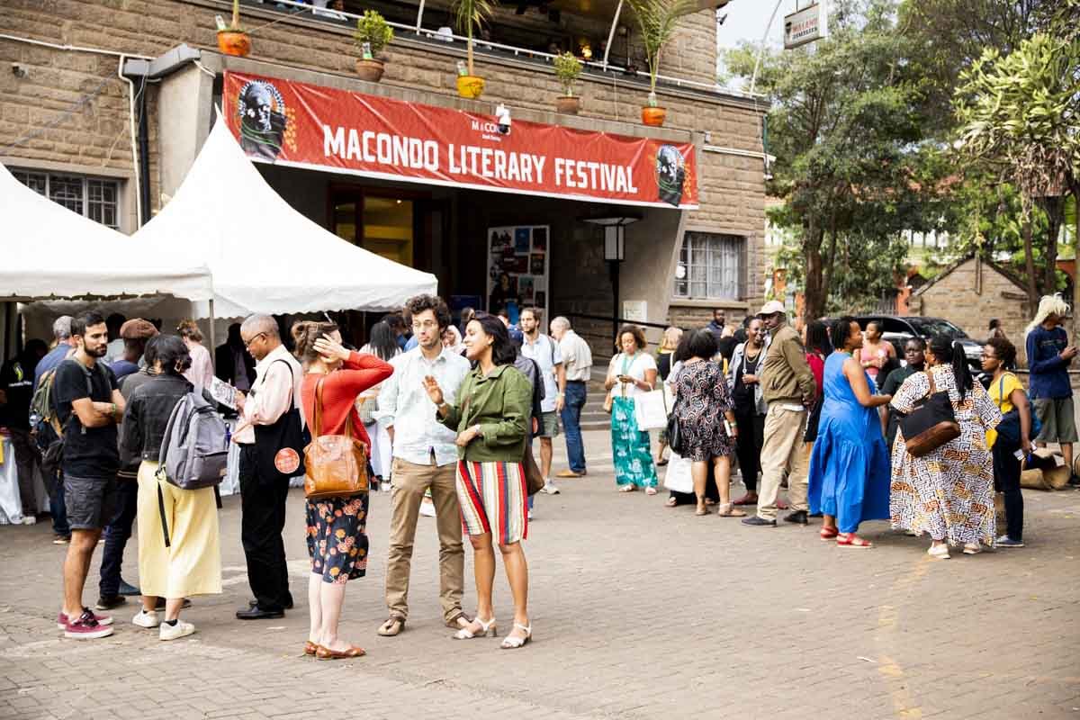 Crowd of people gathered outside under a red banner that reads 'Macondo Literary Festival,' with tents and trees in the background.