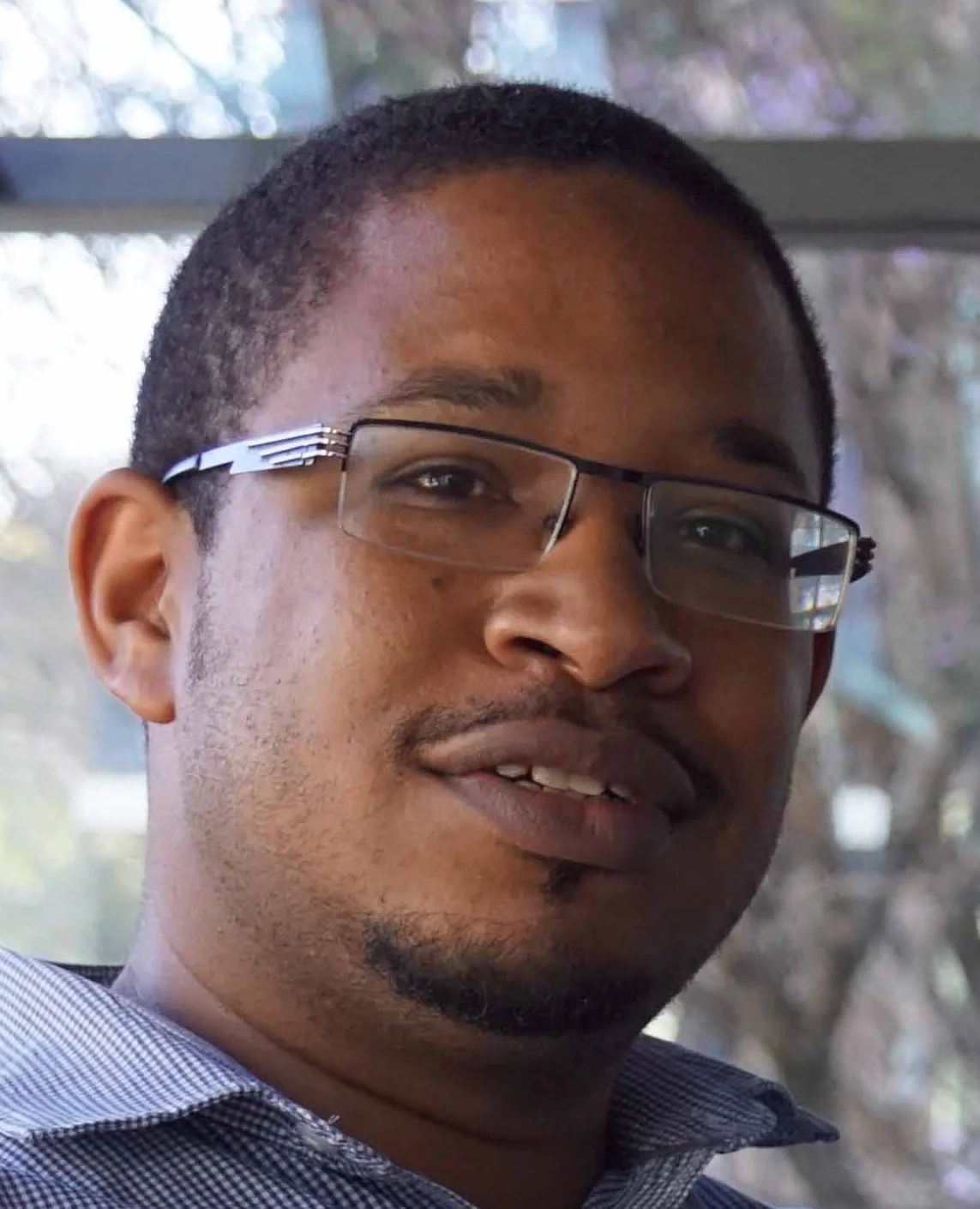 A man with glasses and a short beard smiling indoors with a window and trees outside in the background.
