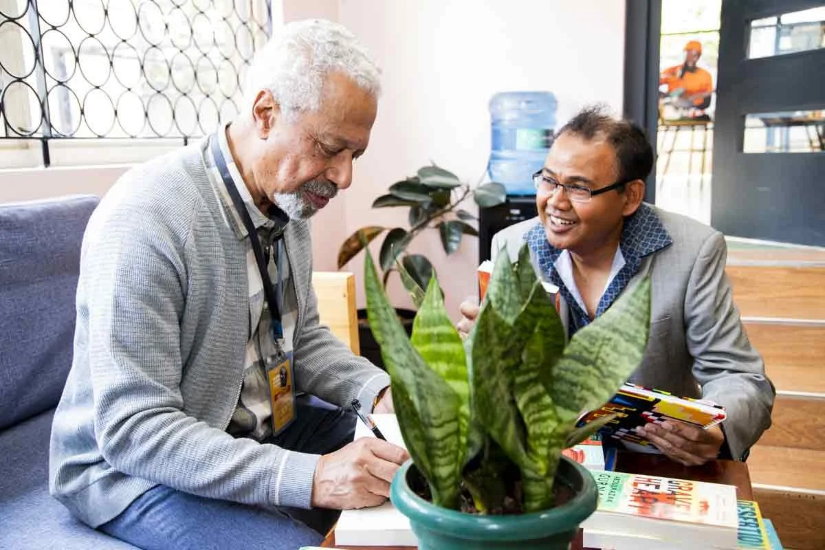 Two men having a friendly discussion in an office or waiting area, with a green potted plant in the foreground and a water cooler in the background. One man with white hair and a gray cardigan is writing or signing something, while the other man with
