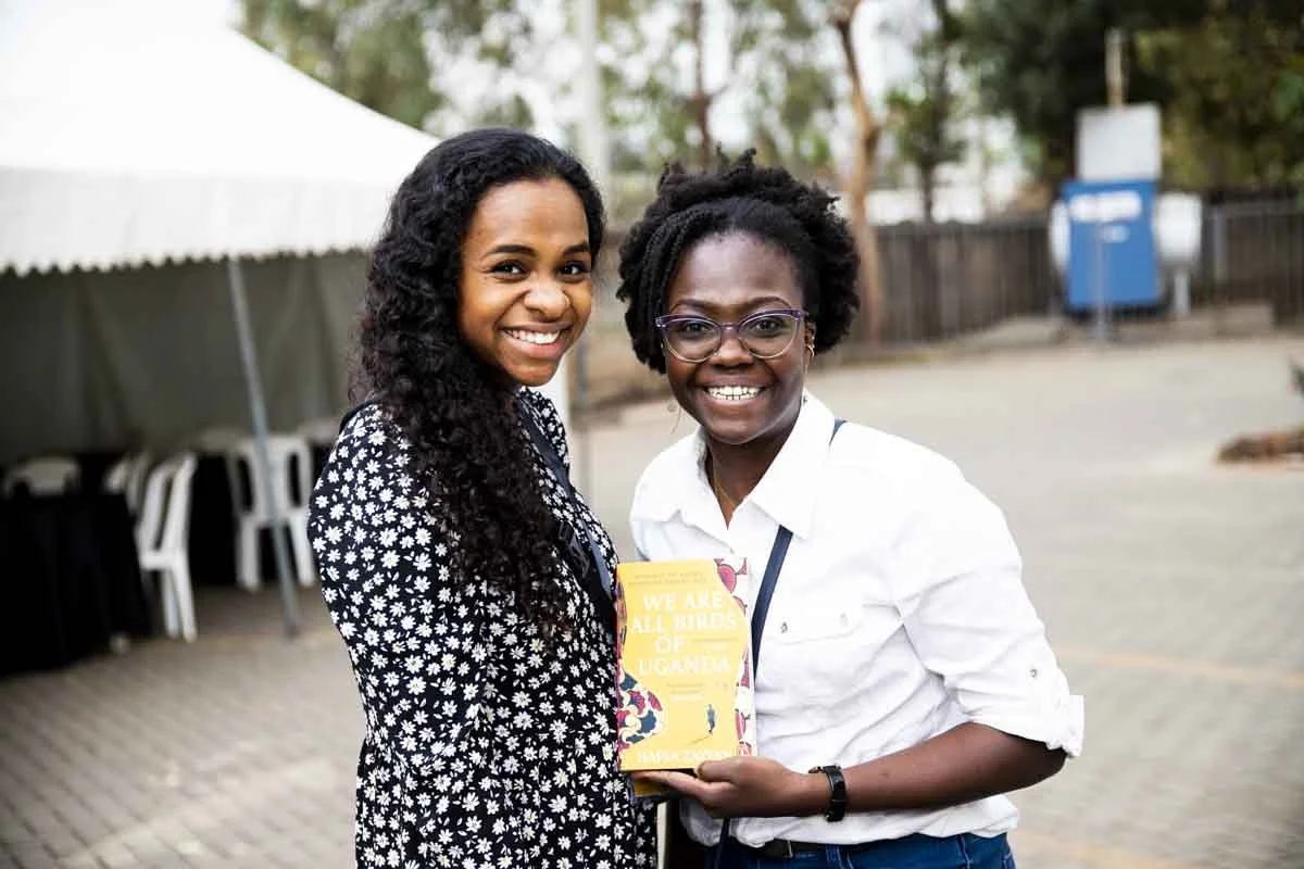 Two smiling women standing outdoors, one holding a yellow book titled "We Are All Refugees of Uganda".