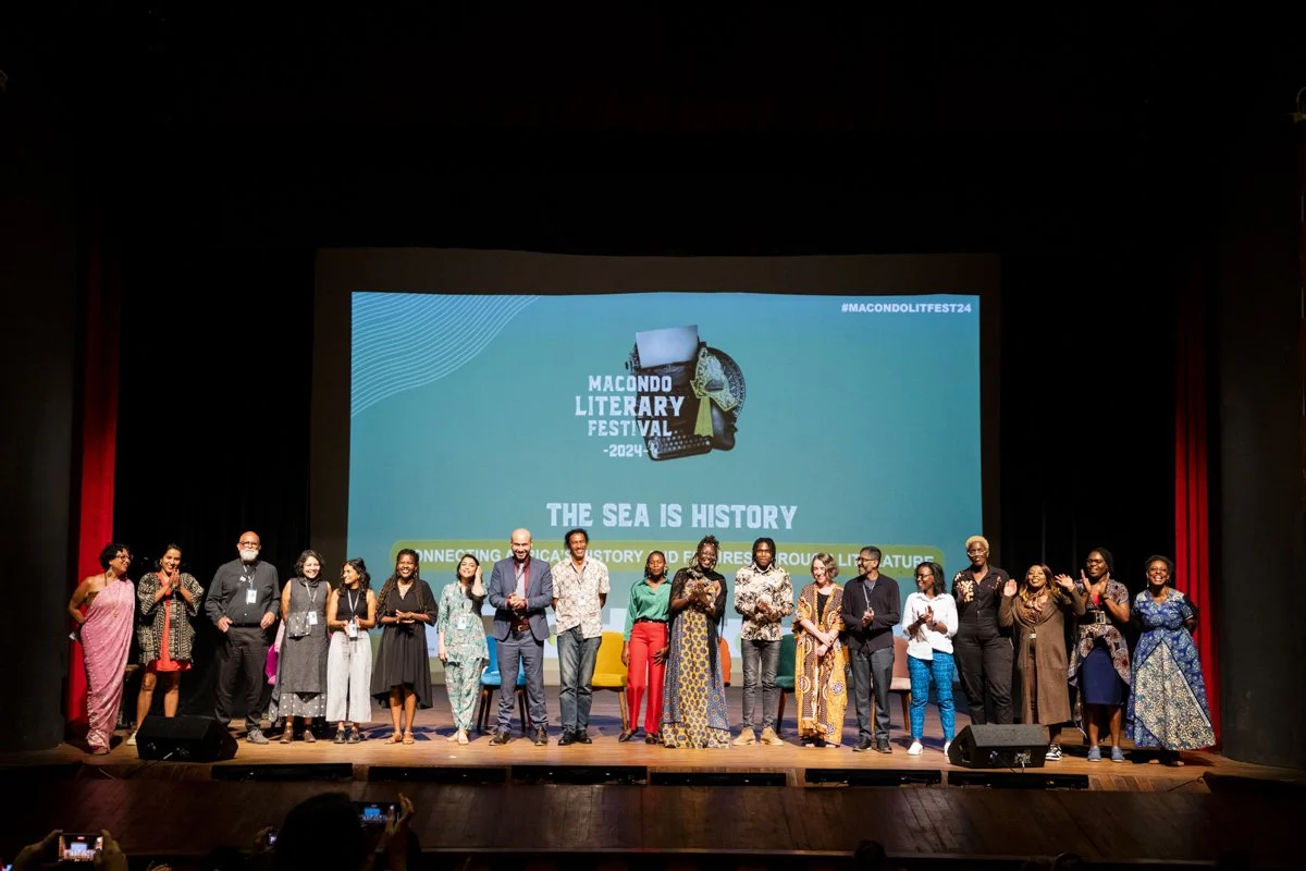 A group of approximately twenty people standing on a stage at the Macondo Literary Festival 2024. They are posing for a photo in front of a large blue screen with festival branding and the slogan "The Sea Is History." The stage has red curtains on ea
