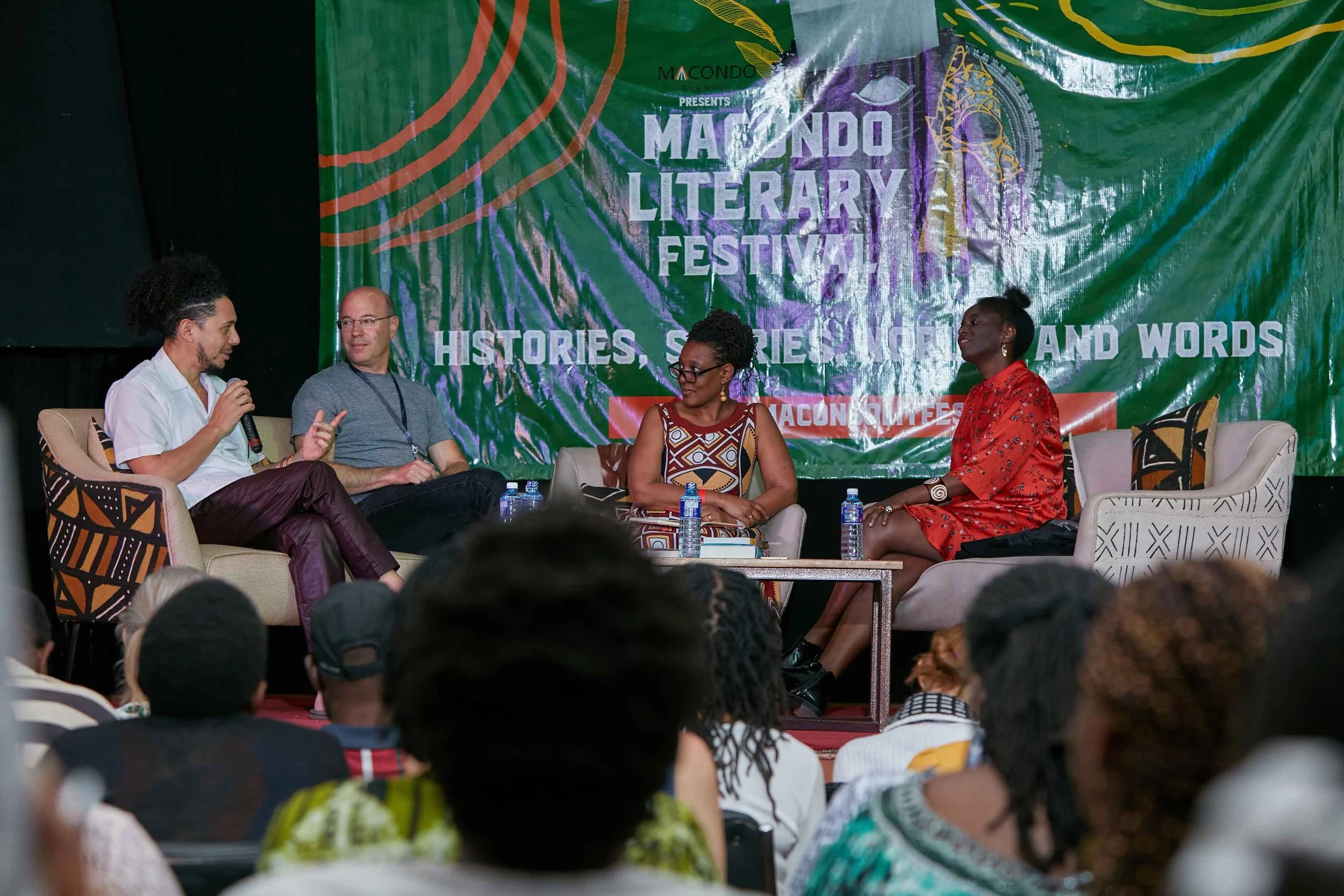 Four people participating in a panel discussion at the Macondo Literary Festival, with a green and purple backdrop that reads 'Macondo Literary Festival' and 'Stories, Stories, Voices, and Words.' There are an audience in front of them.
