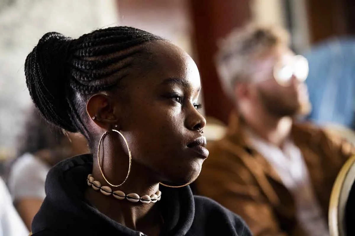 A young woman with braided hair, wearing large hoop earrings and shell necklace, attentively listening in a setting with other blurred attendees.