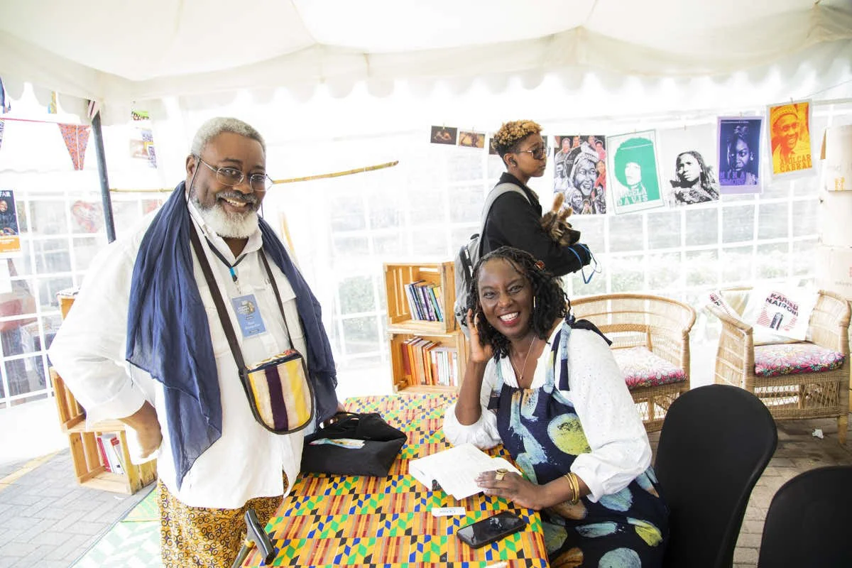 Three people inside a tent at a book fair, with two women seated at a table and a man standing nearby. The woman on the right is smiling and talking on a phone, with papers and a phone on the table. The man on the left is smiling at the camera, weari