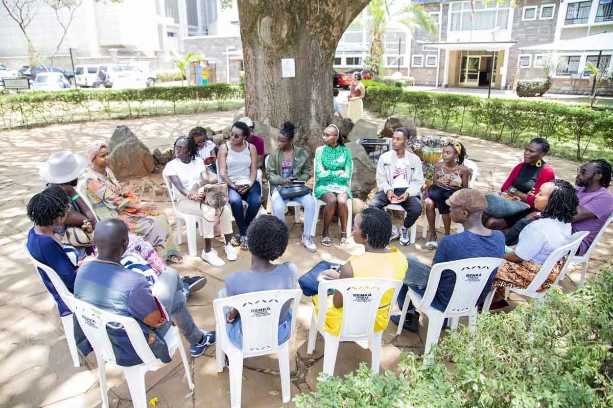 A group of people sitting in a circle outdoors under a large tree, participating in a discussion or meeting, with additional people standing in the background near a residential building.