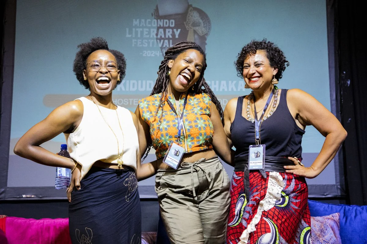 Three women smiling and posing for a photo on stage at the Macondo Literary Festival 2024, with a screen behind them displaying festival information.