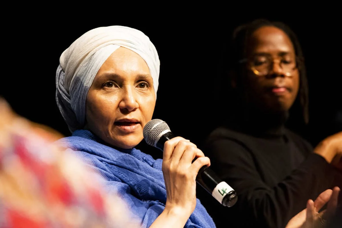 A woman wearing a beige headscarf and blue shawl speaking into a microphone on stage, with a person wearing glasses in the background.